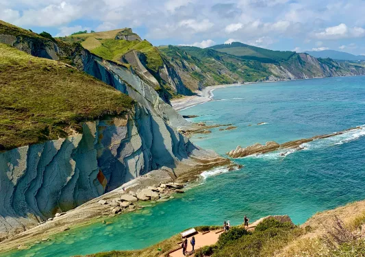Wide shot of rocky coastline. Blue water, layered rocks, guests in distance.