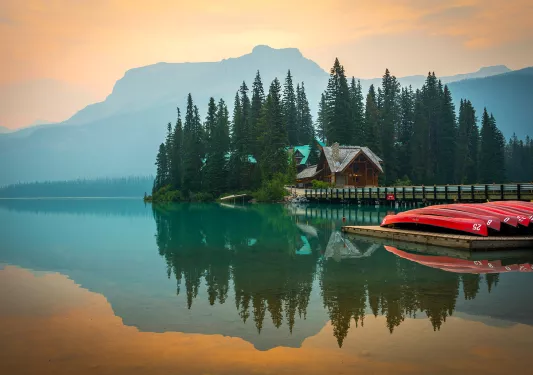 Wide shot of large lake, wooden house, trees, canoes, mountains visible.