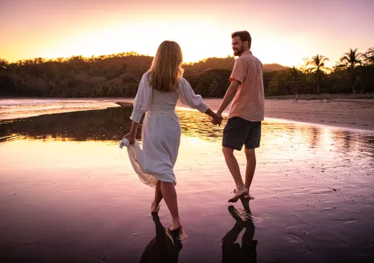 Two guests holding hands, walking along shore during sunset.