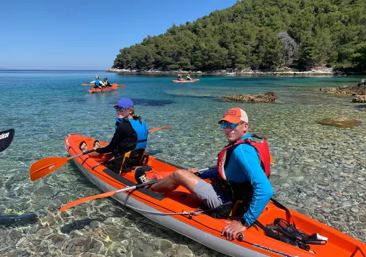 Guests pushing off to kayak, looking at camera, coastal forest in distance.