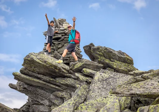 Two guests on craggy clifftop, hanging onto constructed rock pile.