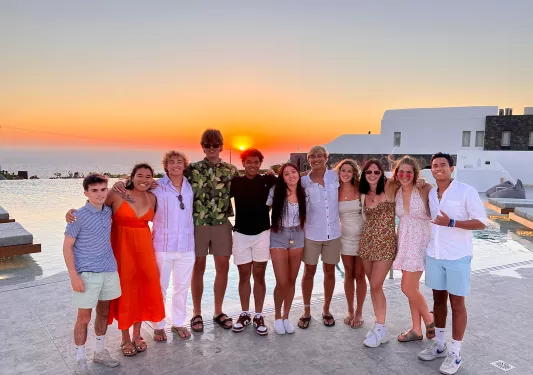 Group of young guests, arms over each other, sunset, white building behind them.