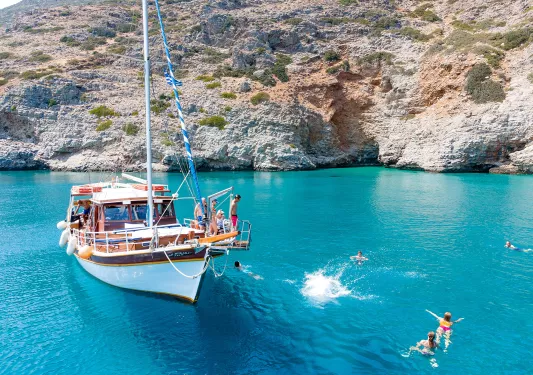 Guests near and on boat, swimming, relaxing, rocky coast nearby.