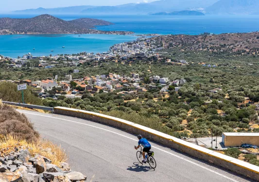 Guest cycling on road, arid coastal town in distance, ocean behind it.