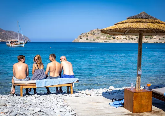 Four guests on rocky beach sitting on bench, overlooking ocean, one looking back at camera.