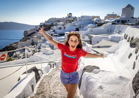Guest emoting to camera, white walled coastal buildings behind her.