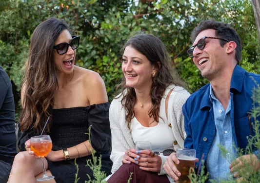 Three guests laughing together, glasses in hand, trees behind them.