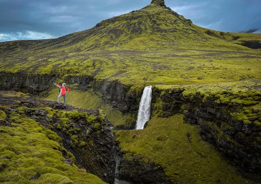 Guest in hiking gear amidst green mountainside, peak, waterfall behind her.