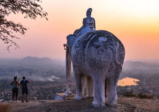 Two people standing beside a large elephant statue as the sun sets in India