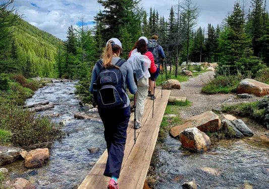 Three guests hiking over stream, forest, clouds, hillside in distance.