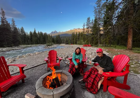 Two guests in red chairs sitting around fire, river, trees, mountain background.