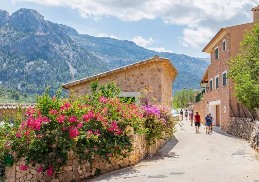 Guests walking through mountain village w/ tan stone houses, hills in distance.
