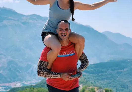 Two guests on hilltop, one on the others shoulders, mountains behind them.