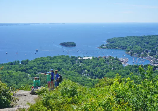 Wide shot of a coastal vista, sailboats visible in water, group of young guests in foreground.