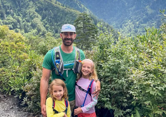 Father and two daughters smiling for camera, large forest and mountain in background.