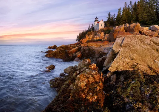 Wide shot of craggy inlet, small white lighthouse, sunset.