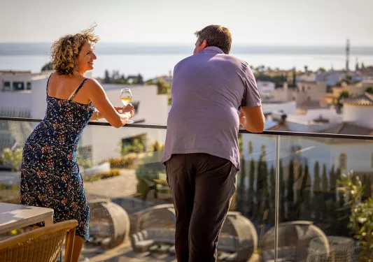 Two guests standing on a balcony overlooking a town on the sea