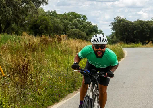 Guest on road cycling towards camera, smiling, trees, clouds behind.