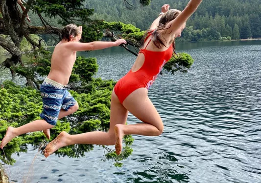 Two young guests jumping into ocean, fog, hills behind them.