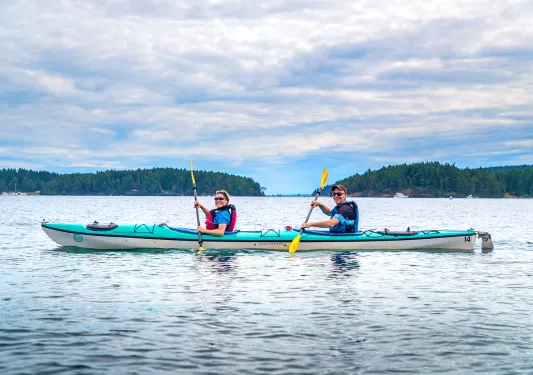 Two guests kayaking, smiling at camera, hills, sky behind them.