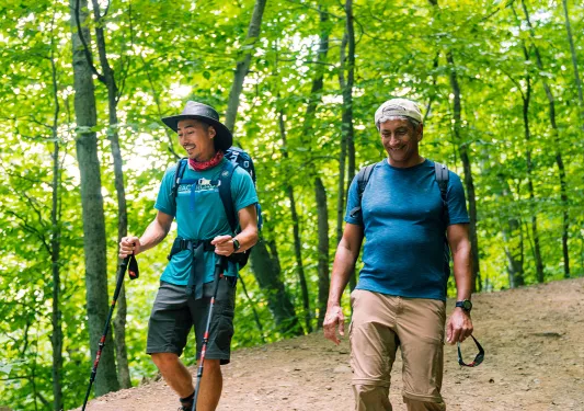 Two guests with hiking gear walking down forest trail.