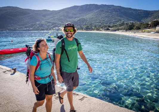 Two guests walking on stone pier, ocean to their left, beach and hills in background.