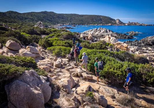 Eight guests hiking on rocky shore, ocean to their right, hills in distance.