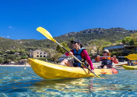 Two guests beginning to kayak on blue water, beach houses in background.