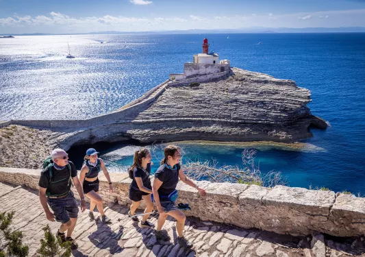 Four guests hiking up rocky staircase, small white lighthouse & ocean in background. 
