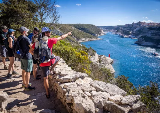 Six guests overlooking blue river, pointing towards rocky cliffs and hillside houses.