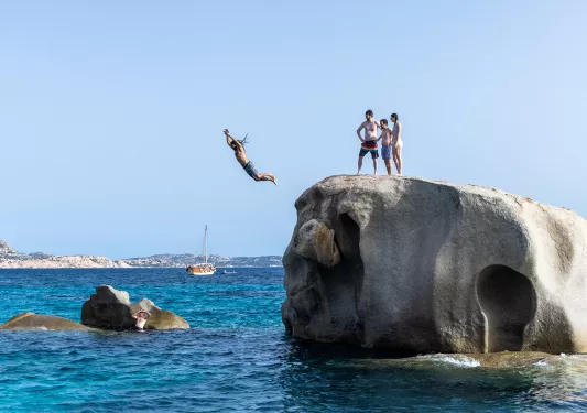 Four guests jumping off large rock, one in the water below.