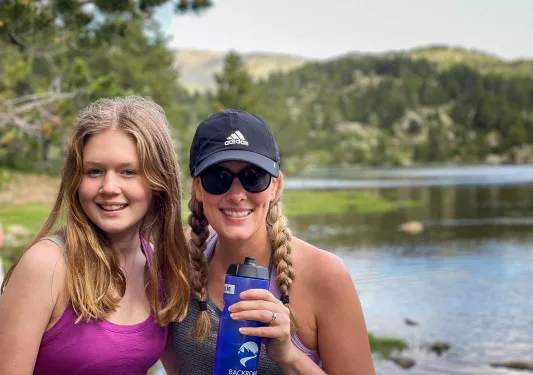 Two women smiling at the camera while hiking beside a lake