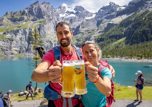 Two guests w/ beer in front of lake, mountains in background.