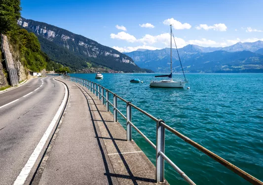 Wide shot of coastal road, blue water, cliffside, small boats.