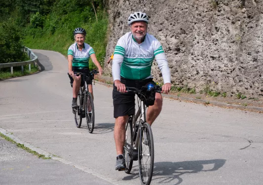 Two guests biking down road, Backroads jerseys on, smiling.