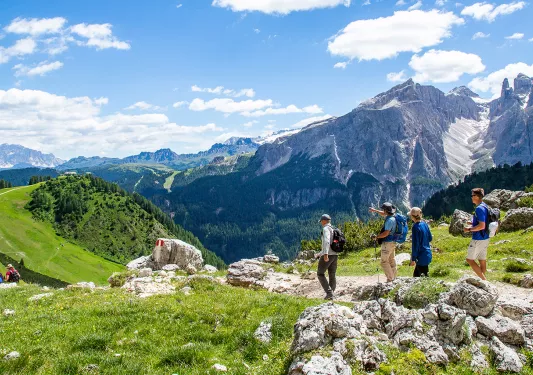 Group of guests hiking down mountain trail, Dolomite range in background.