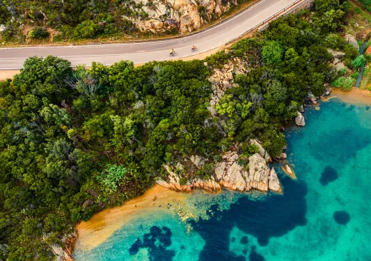 Overhead shot of guests cycling on coastal road, small beach, blue water.