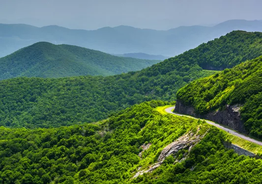 Wide shot of mountain road, overlooking large forested mountains.