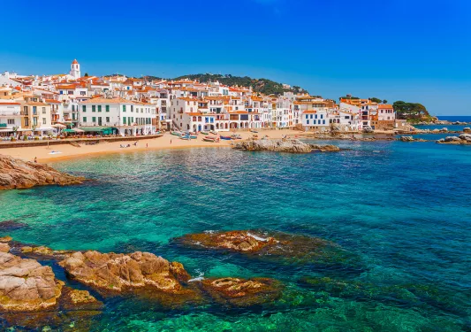 Shot of Costa Brava coastline, blue water, tan and white buildings.