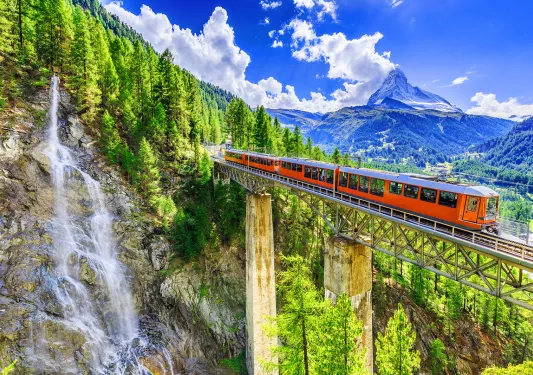 Wide shot of mountain train passing bridge, forest, mountains, sky in distance.