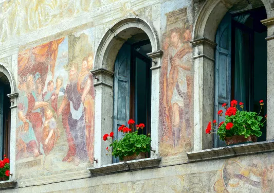 Shot of ancient mural on side of building, open windows, flowers.