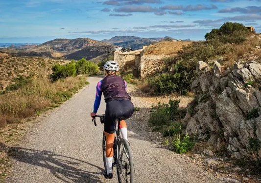 Guest cycling down gravel road, towards stone ruins, golden hills. 