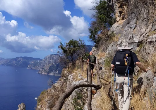Two guests hiking along rocky cliff, flimsy wooden fence on their left.