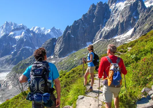 Three guests on mountain trail, looking towards craggy cliff faces in distance. 