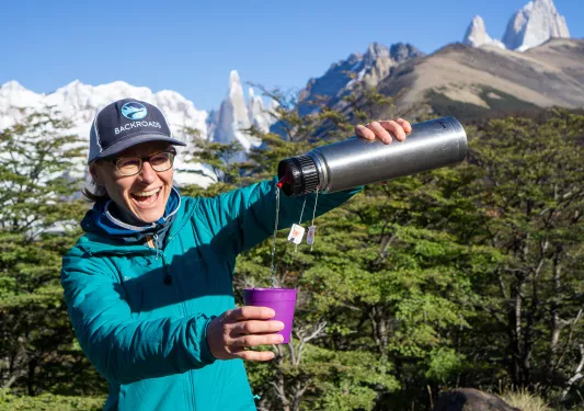 Leader pouring water into yerba mate infuser, trees, snowy peaks behind them.