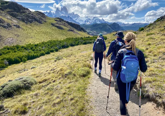 Three guests walking down meadow, valley, hills, clouds in distance. 