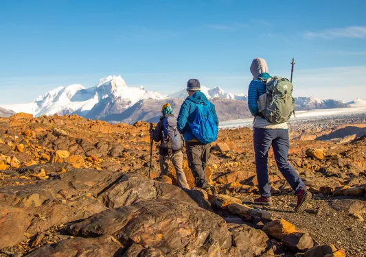 Three guests walking over orange rocks, snowy peaks in distance.