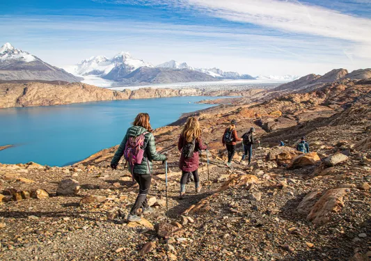 Group of guests walking towards blue lake, snowy mountains in distance. 
