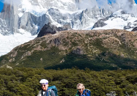 Two guests hiking over small bridge, sharp snowy mountain peaks in distance.