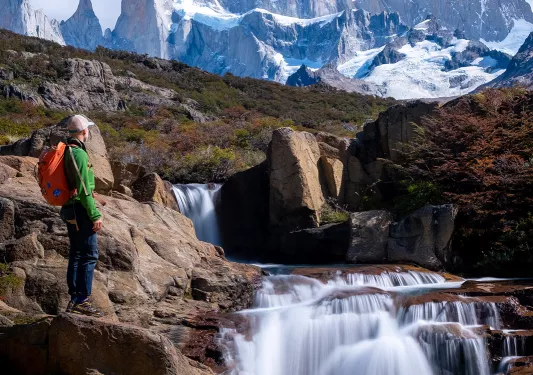 Guest standing next to flowing waterfall, sharp, snowy peaks in background.